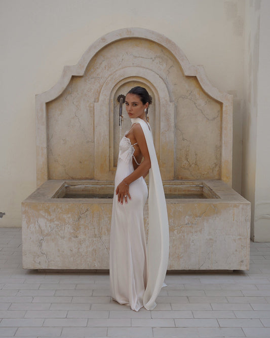 A woman wearing a floor-length ivory or off-white sleeveless gown with a long draped scarf (the Farfalla by Vedere) stands in front of a large stone fountain in a pale beige architectural setting.