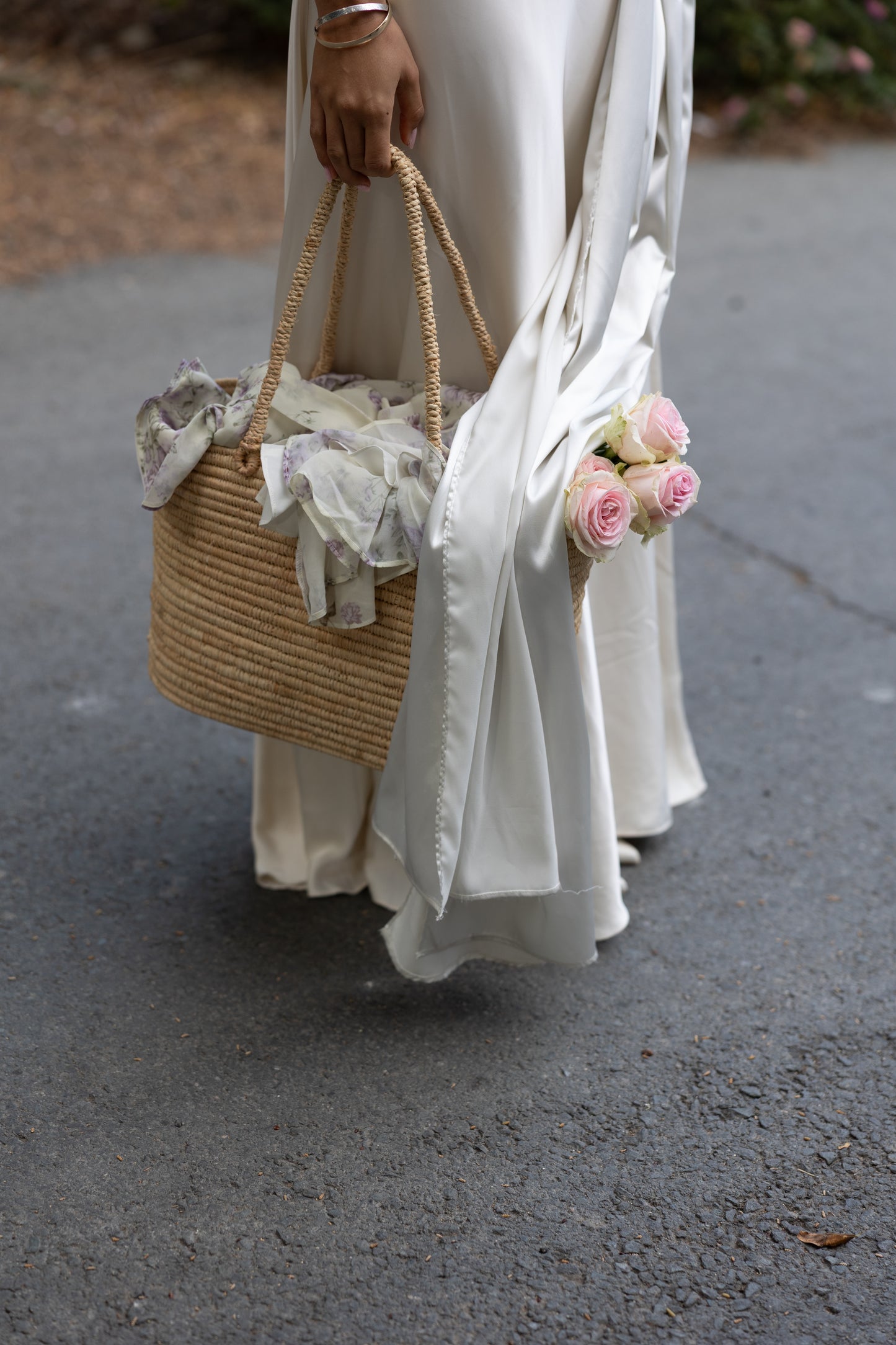 Person holding a woven bag with lace details and pink flowers, wearing a white dress with a white satin scarf draped over the top.