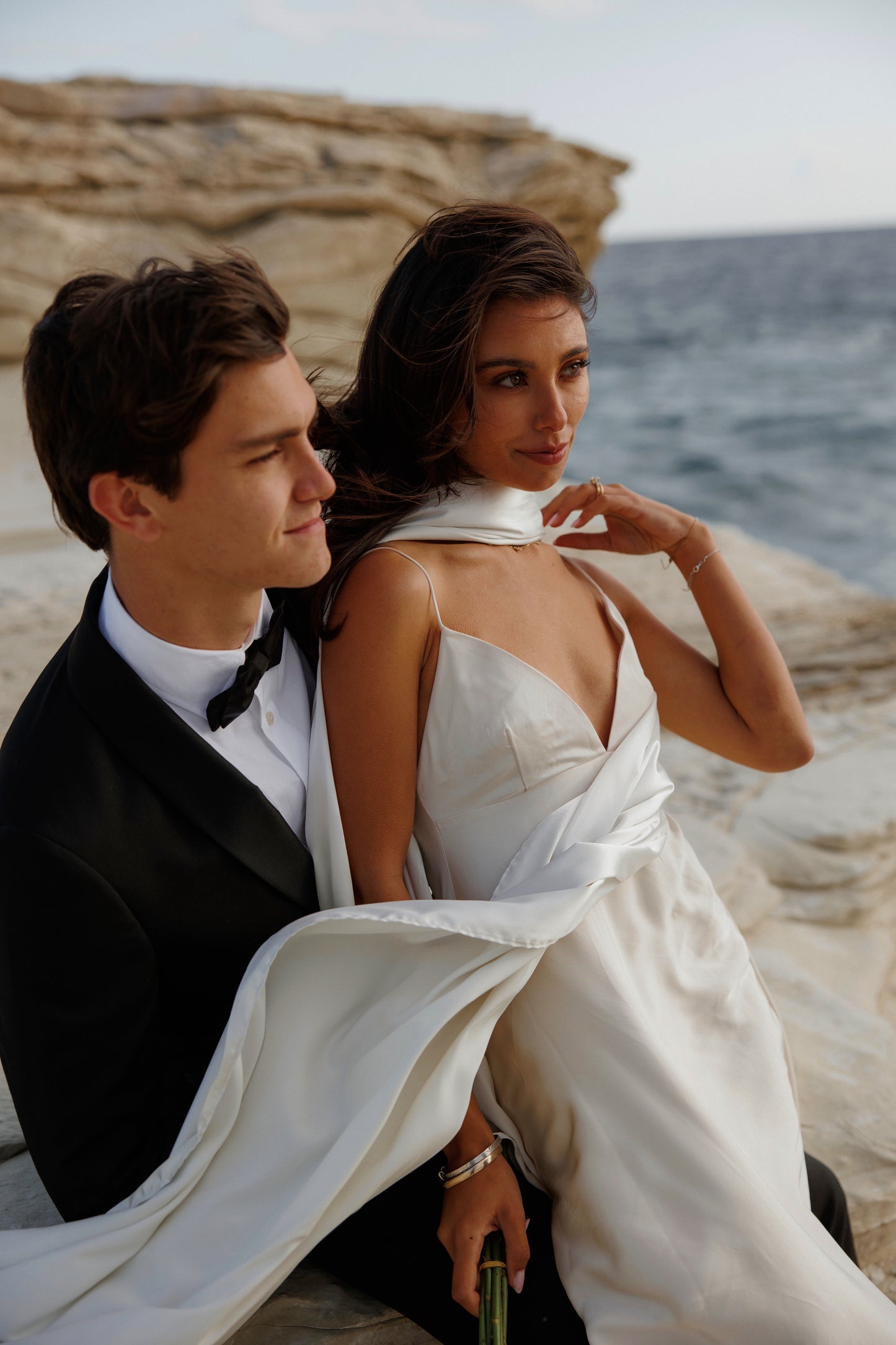 Couple in formal attire sitting on a rocky beach with ocean view. Woman is wearing a long flowy satin scarf.