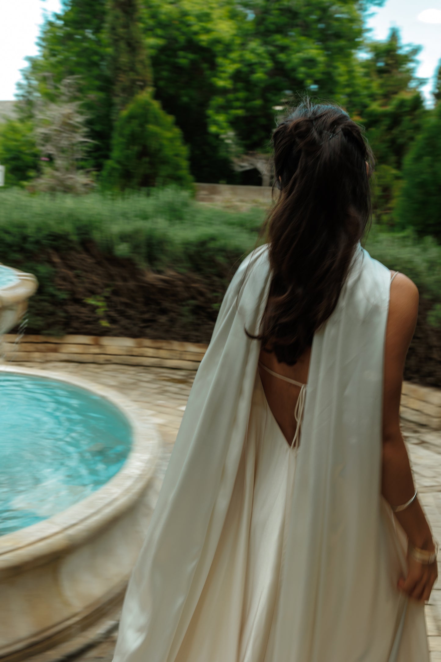 Woman in a white dress and long flowy satin scarf standing by a pool with greenery in the background