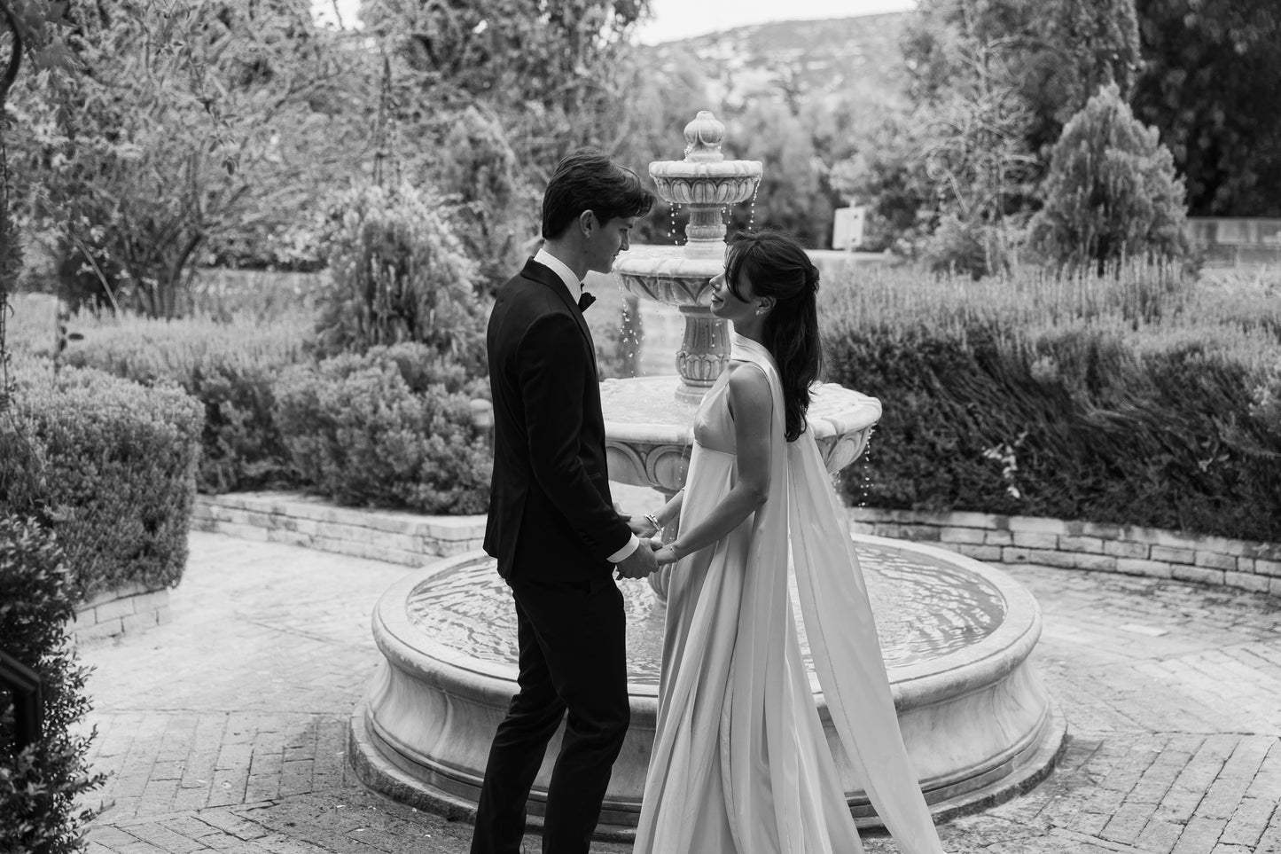 Black and white photo of a couple holding hands in front of a fountain. Bride is wearing long flowy satin neck scarf.
