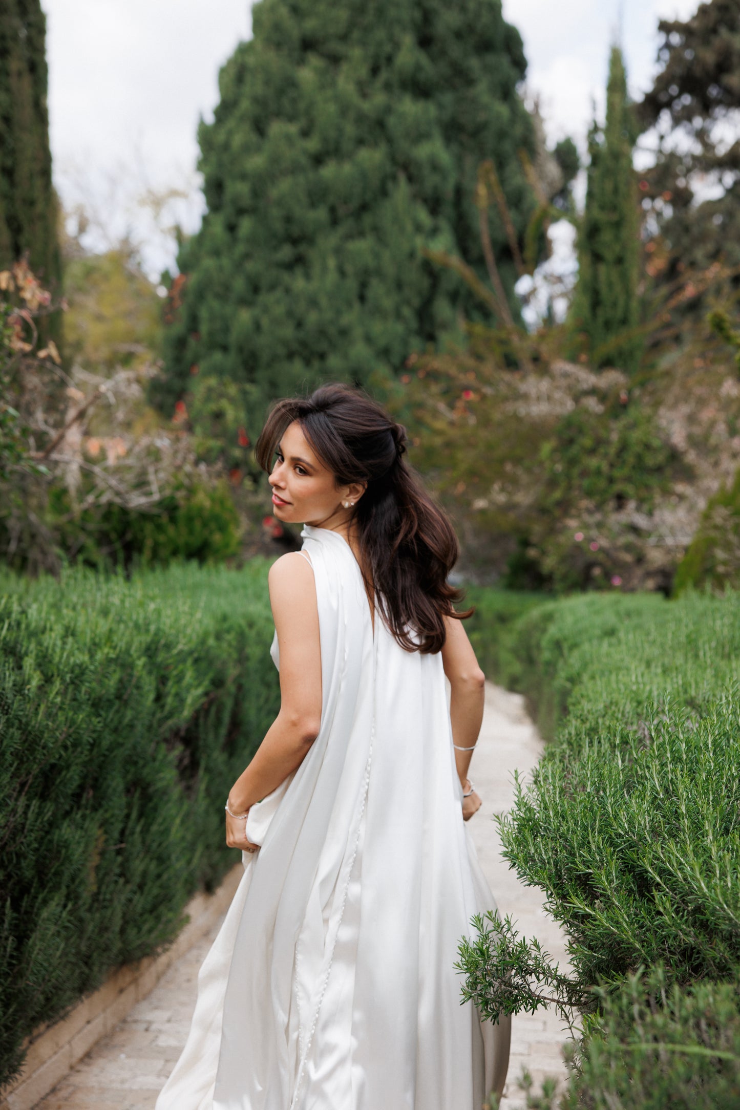 Woman in a white dress walking through a garden path with greenery around wearing a long flowy satin neck scarf.