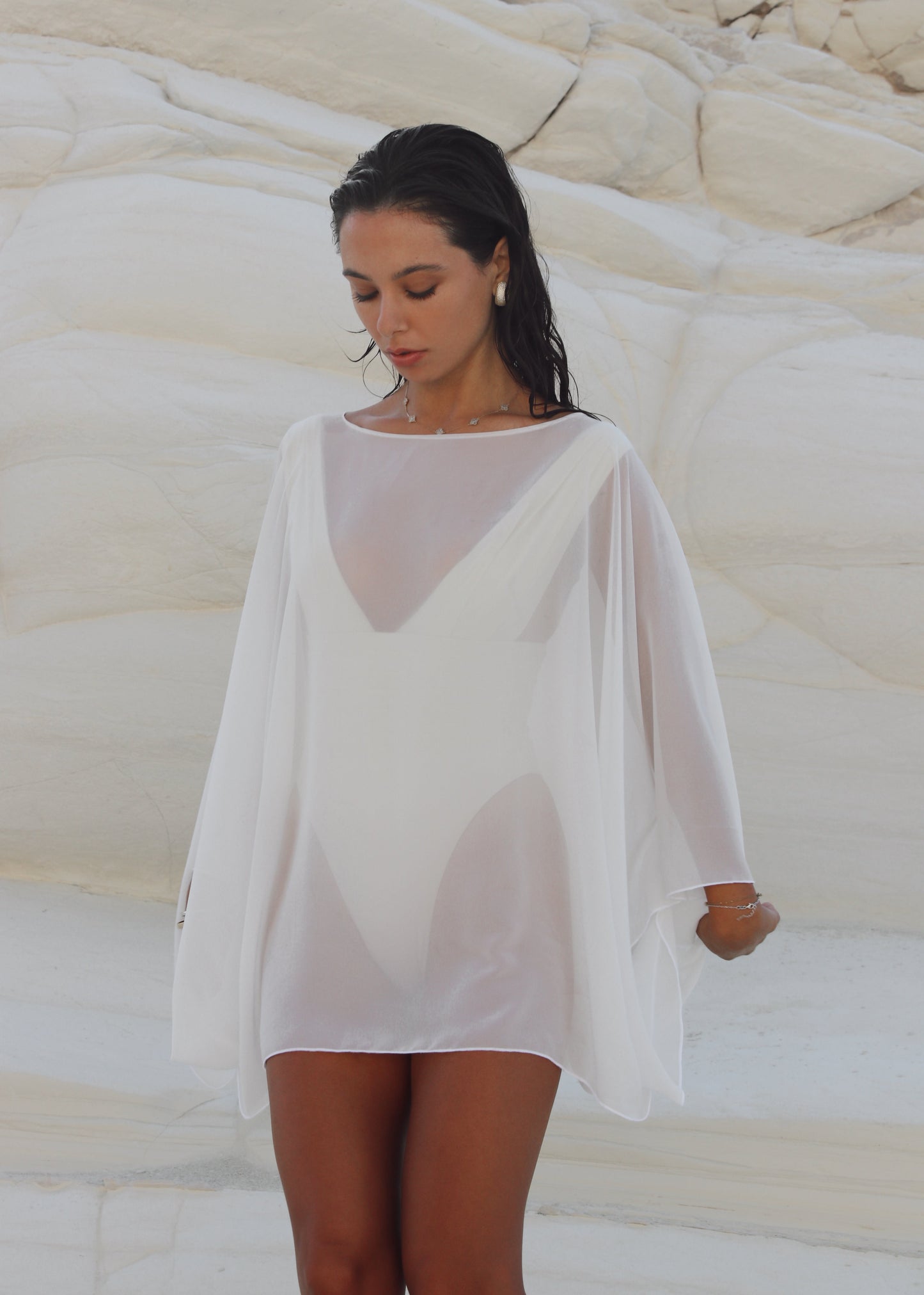 Woman wearing a white poncho with a sheer overlay against a textured beige wall.