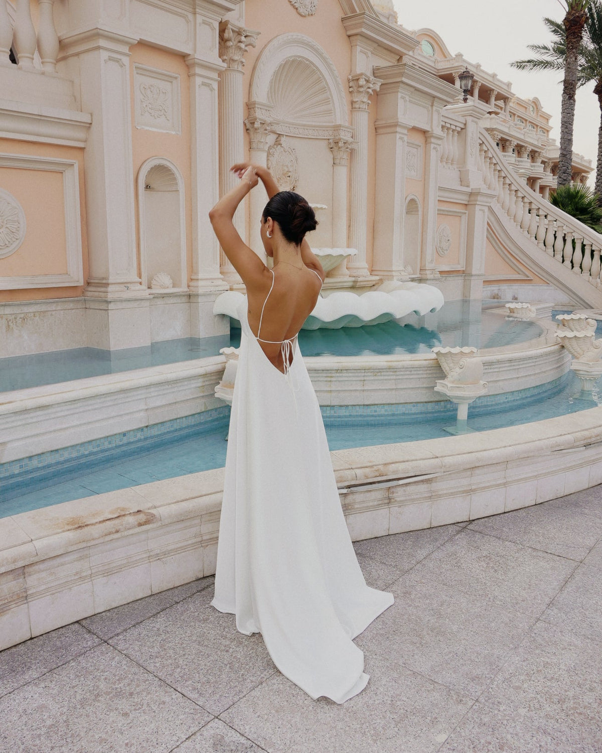 A woman in a flowing white backless gown poses beside a decorative fountain and pale stone building.