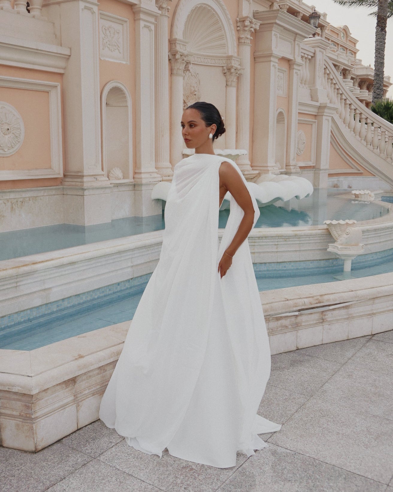 A woman wearing a flowing white draped wedding dress (the Roma Dress and Medusa Cape) stands beside a large outdoor fountain in front of a pale beige architectural building.