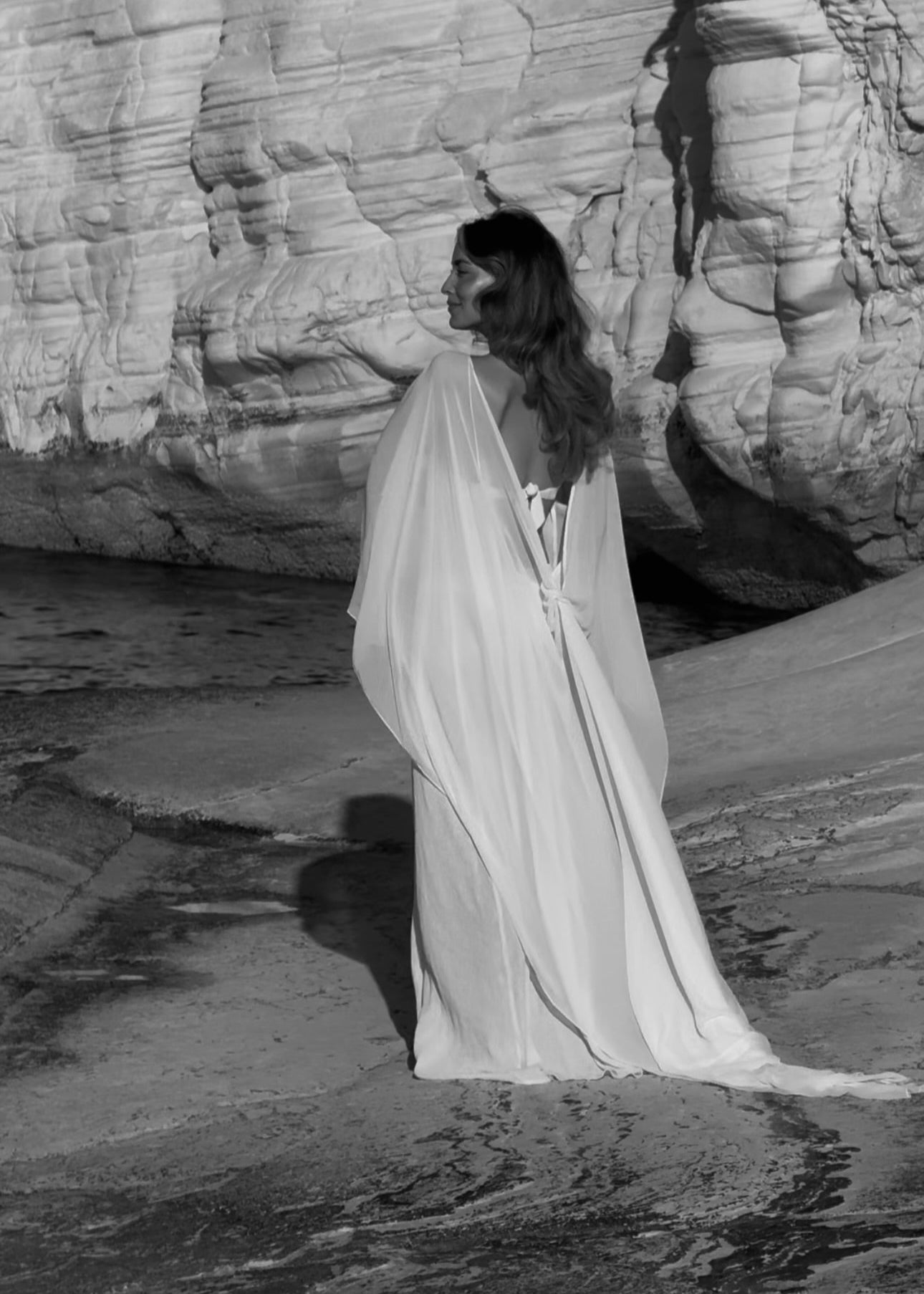 Woman in a flowing white dress standing on a rocky beach with large rock formations.