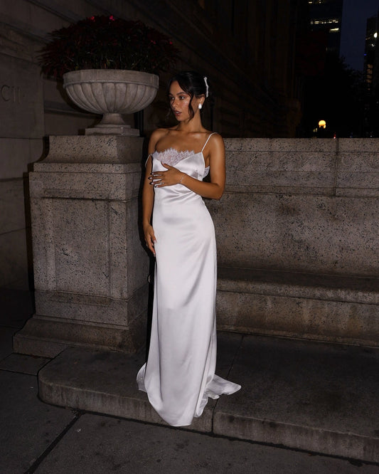 A woman wearing a white satin strapless gown with a sweetheart neckline and lace detailing, standing on stone steps at night.