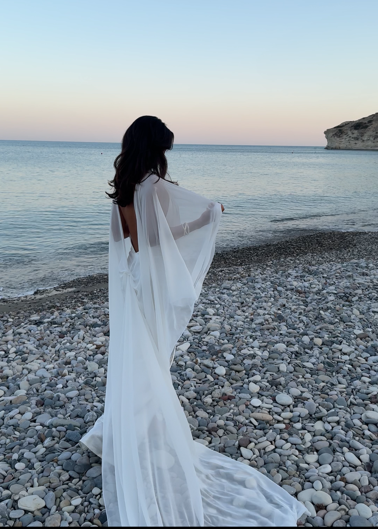 Woman in a white cape standing on a pebbly beach with ocean and sky in the background