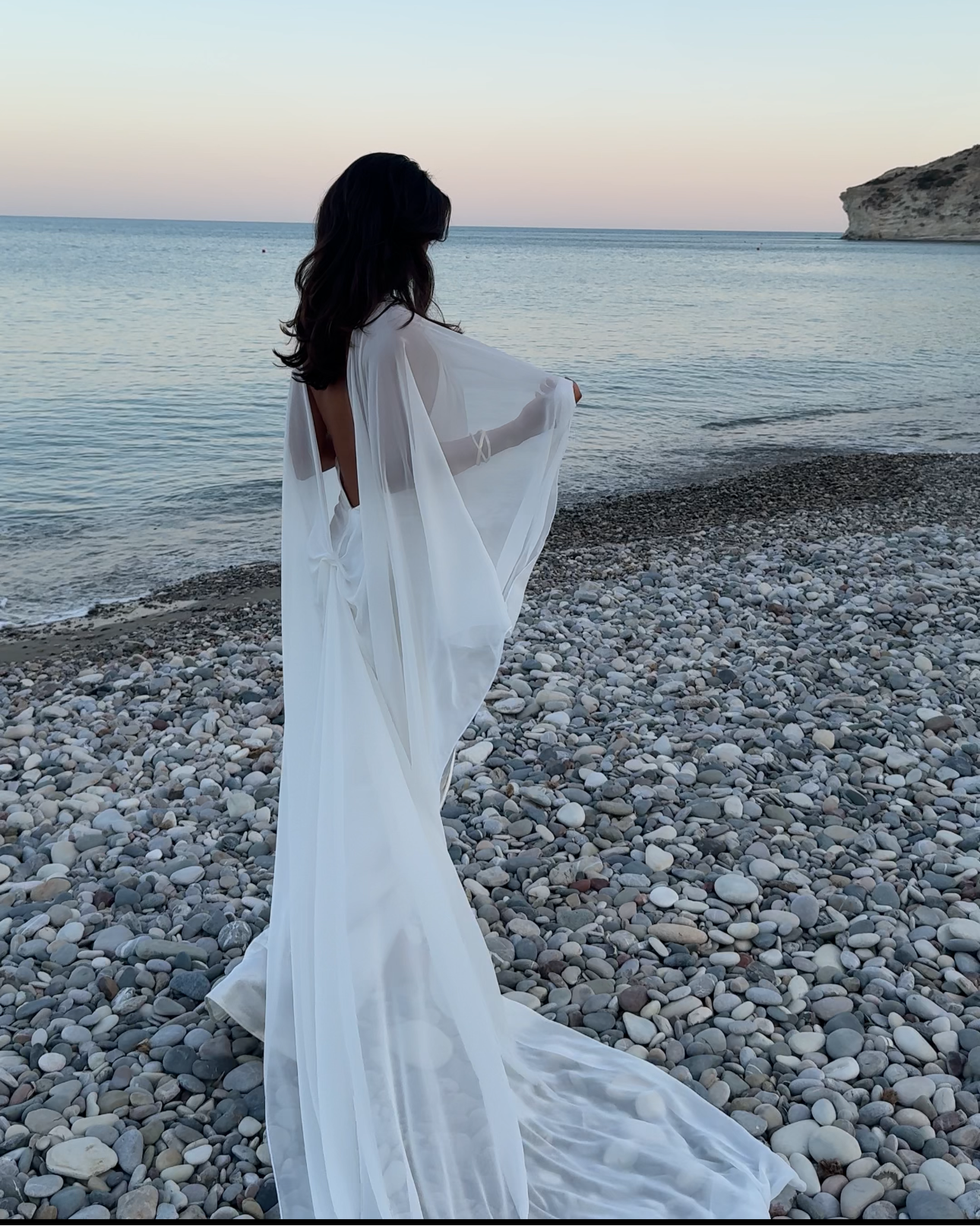 Woman in a white cape standing on a pebbly beach with ocean and sky in the background