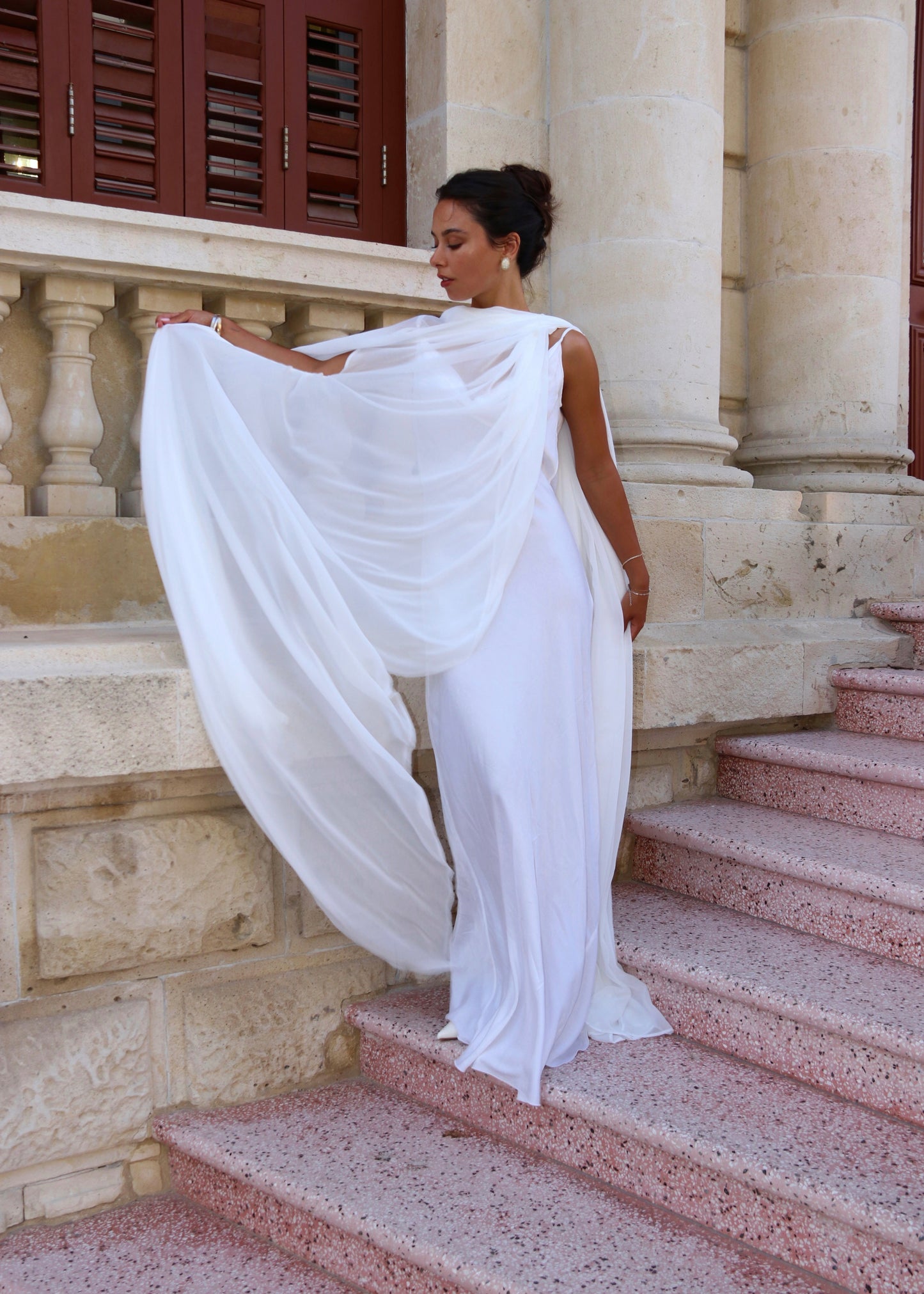 Woman in a white bridal cape standing on stone steps with a building in the background