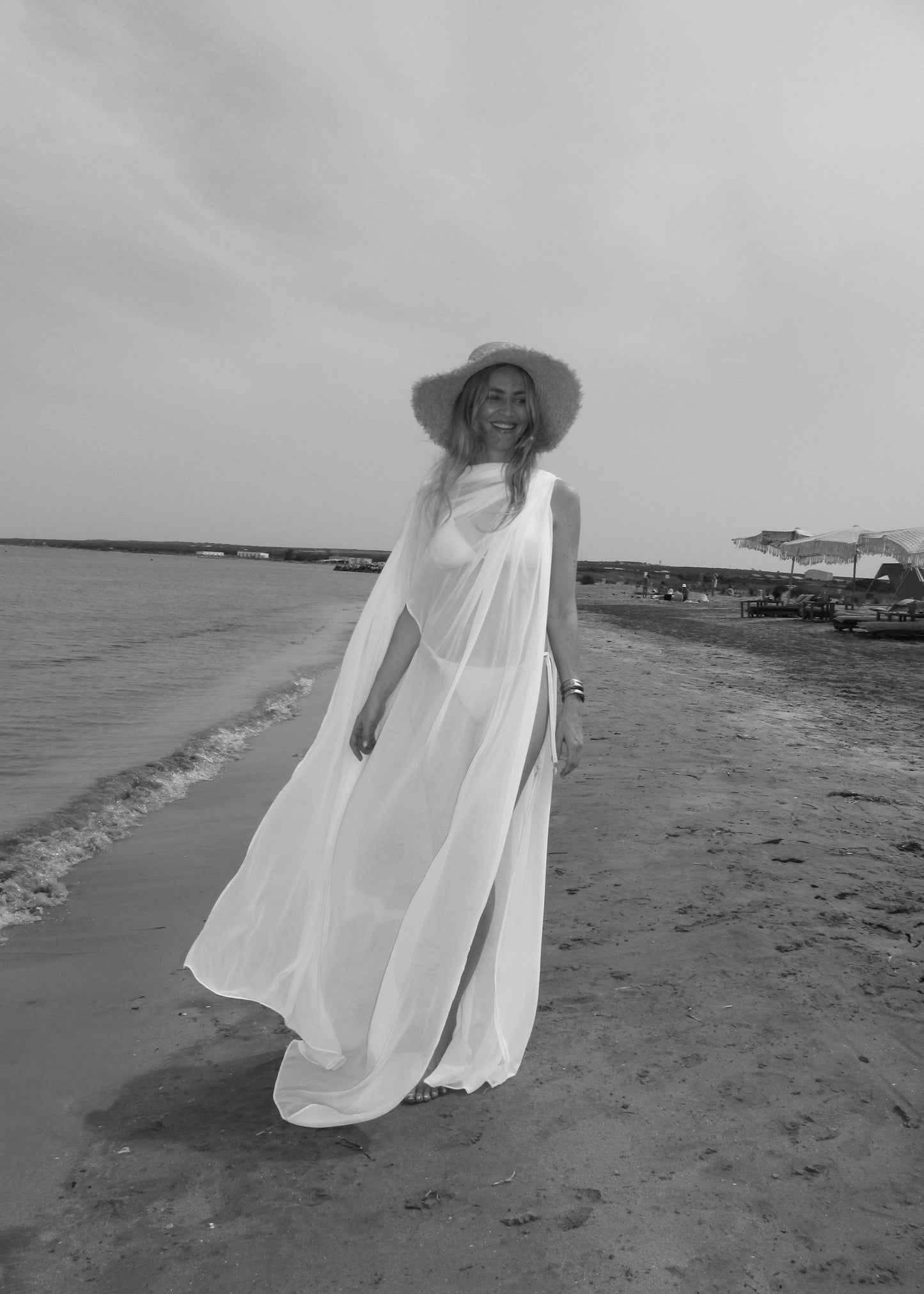 Woman in a long white cape and hat standing on a beach.