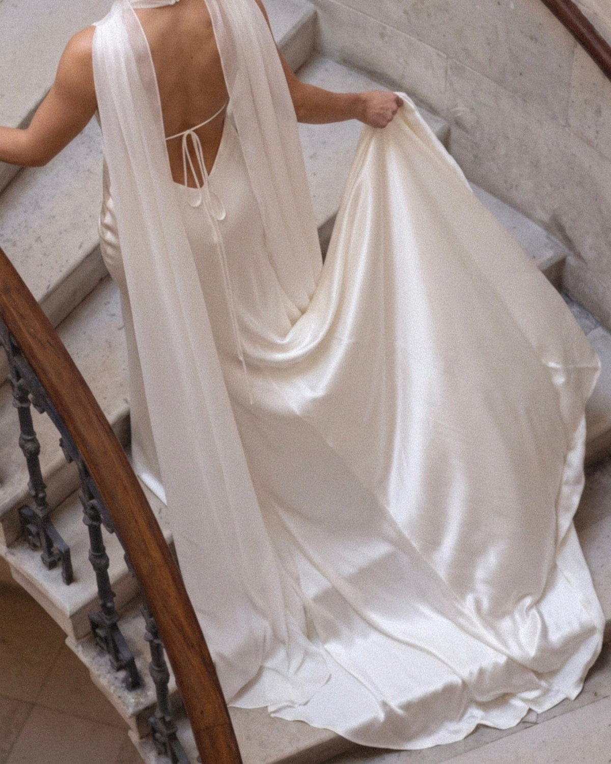 Woman in a white wedding dress and bridal cape standing in front of a fountain in an elegant outdoor setting.