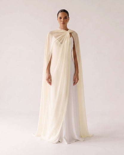 A model stands against a plain light beige studio background wearing a flowing draped gown with a cape-like overlay and a white underlayer.