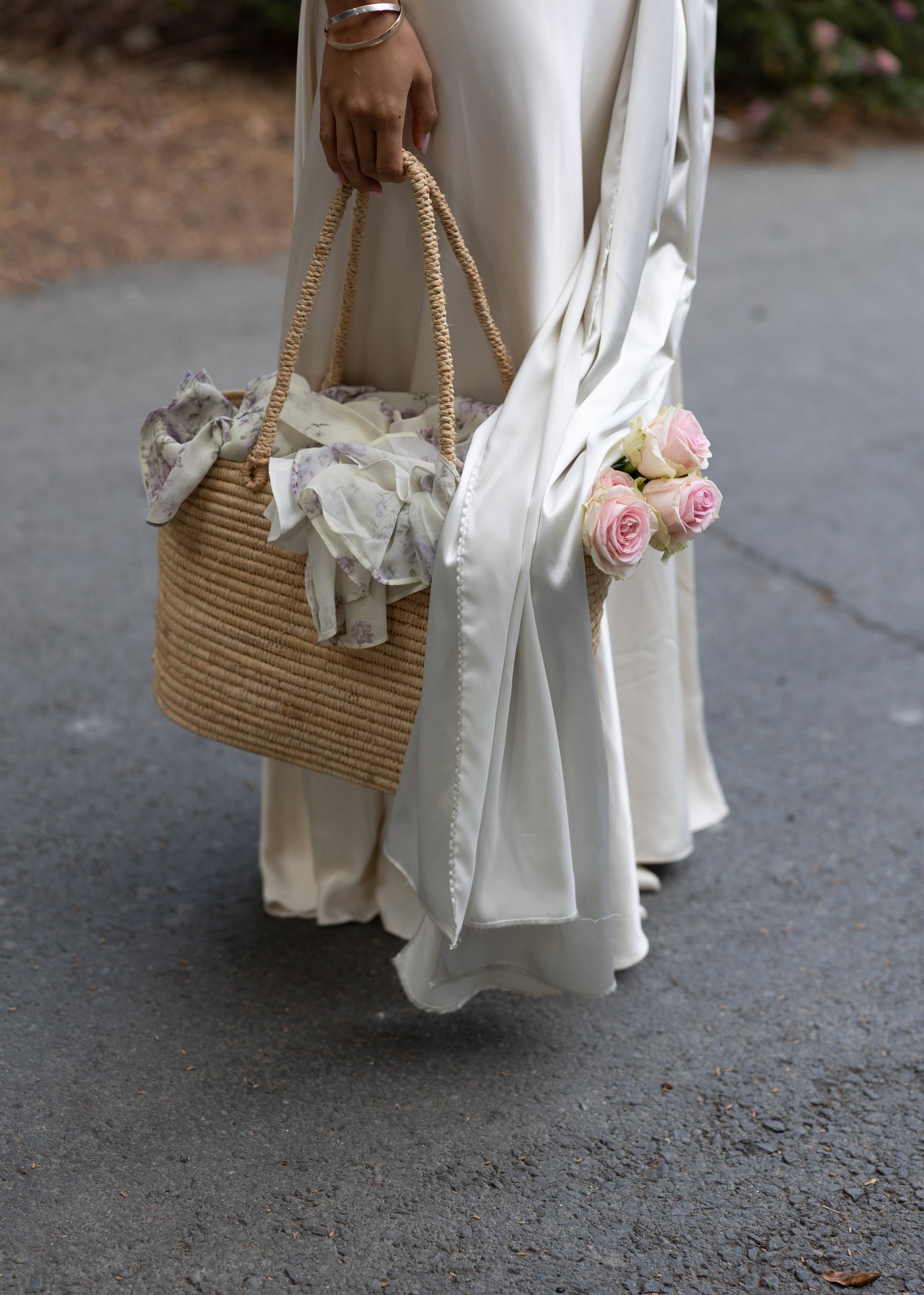 Person holding a woven bag with lace details and pink flowers, wearing a white dress.