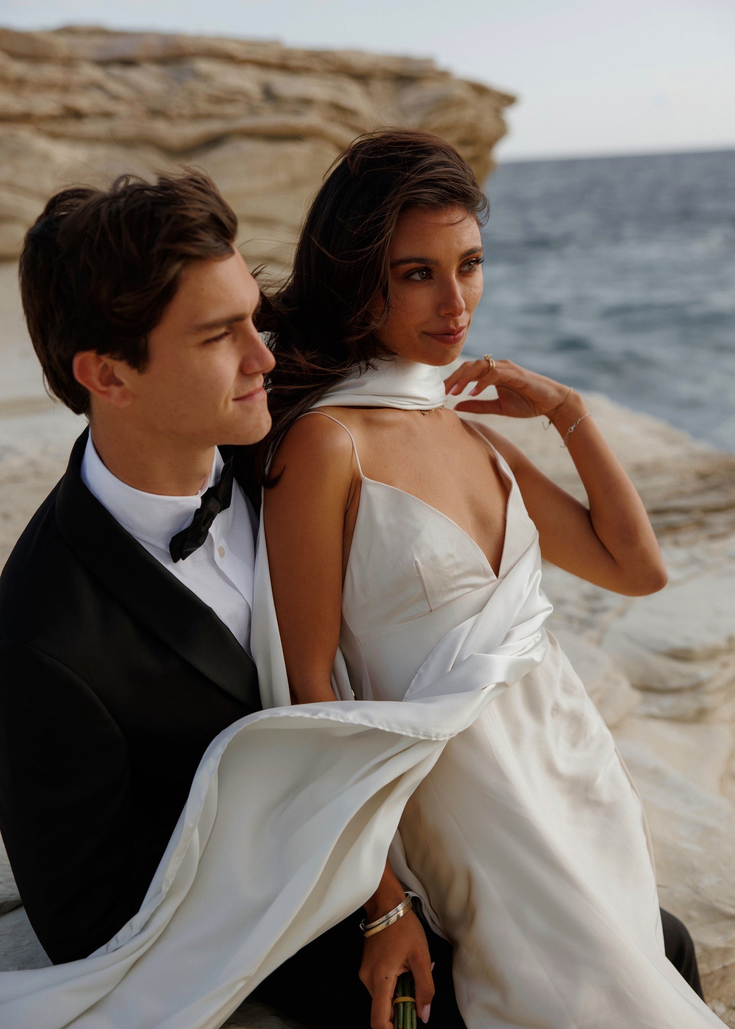 Couple in formal attire sitting on a rocky beach with ocean view