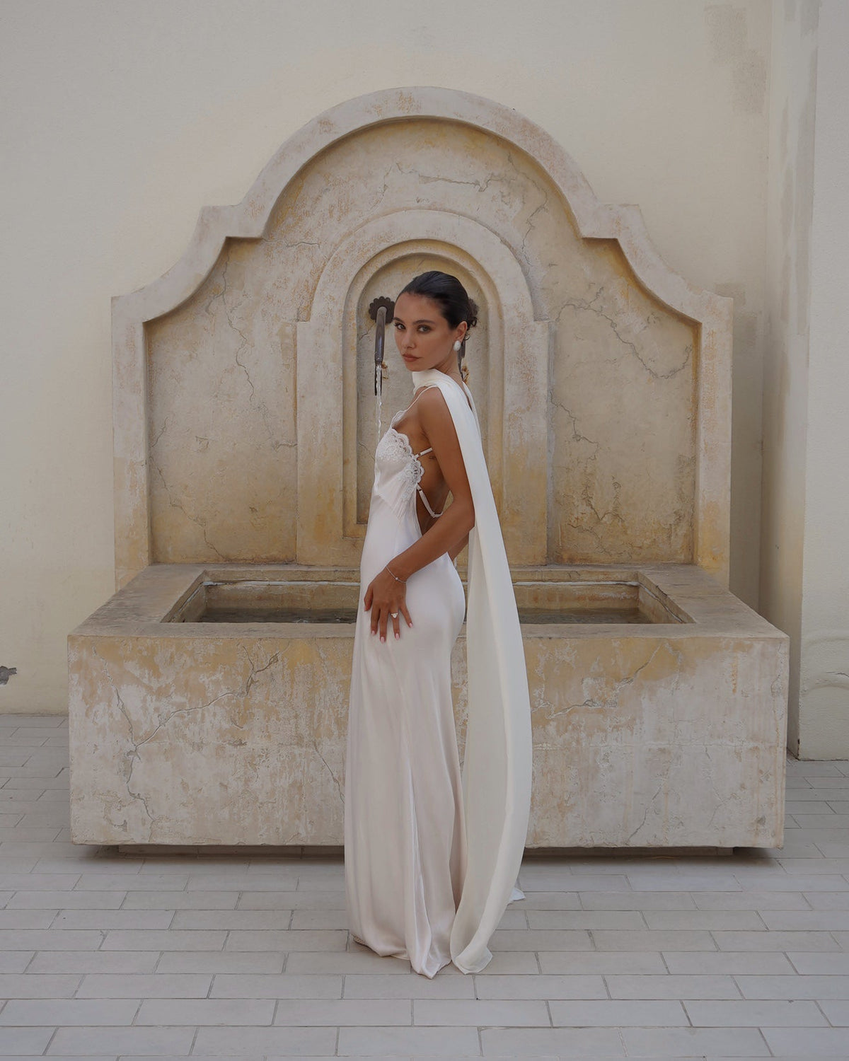A woman wearing a floor-length ivory or off-white sleeveless gown with a long draped scarf (the Farfalla by Vedere) stands in front of a large stone fountain in a pale beige architectural setting.