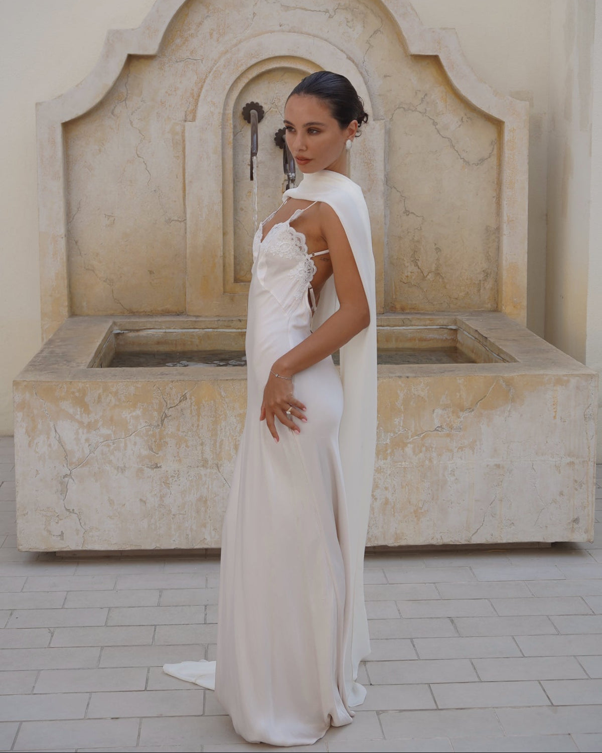 A woman wearing a sleeveless white wedding gown with a draped farfalla scarf by Vedére stands in front of a large stone fountain in a beige courtyard.