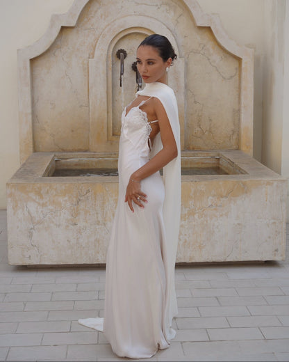 A woman wearing a sleeveless white wedding gown with a draped farfalla scarf by Vedére stands in front of a large stone fountain in a beige courtyard.