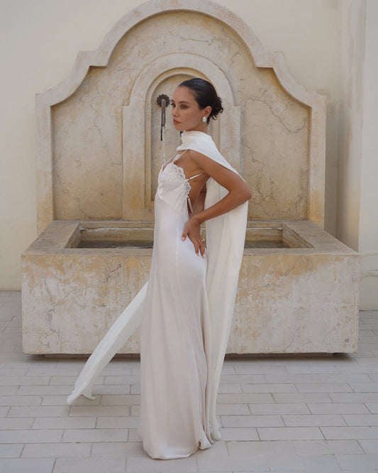 A woman wearing a floor-length white bridal gown with a high neck, sheer draped scarf (Farfalla in Ivory by Vedére) detail, and an open back, standing in front of a large stone fountain.