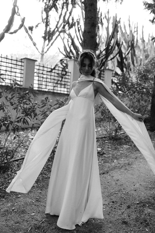 Woman in a flowing dress and white satin scarf standing outdoors with trees and a fence in the background