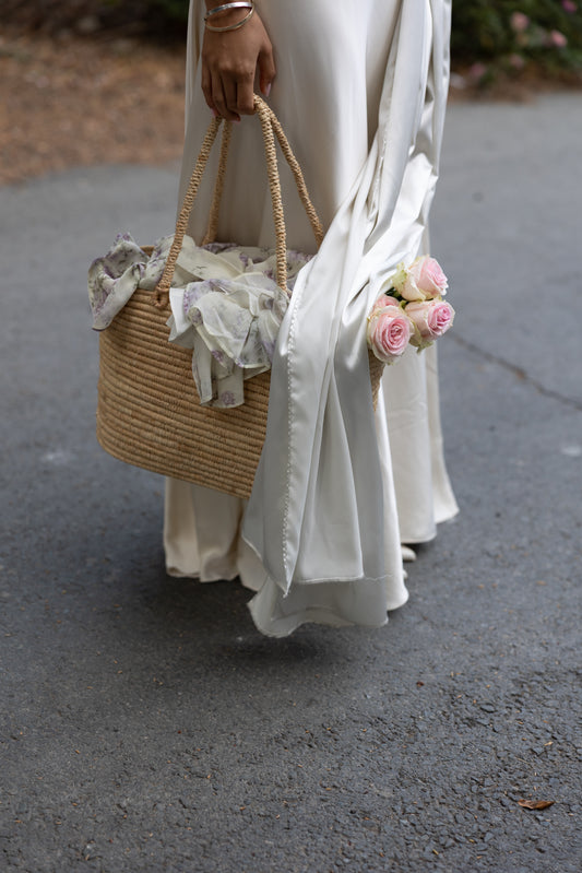 Person holding a woven bag with lace details and pink flowers, wearing a white dress with a white satin scarf draped over the top.