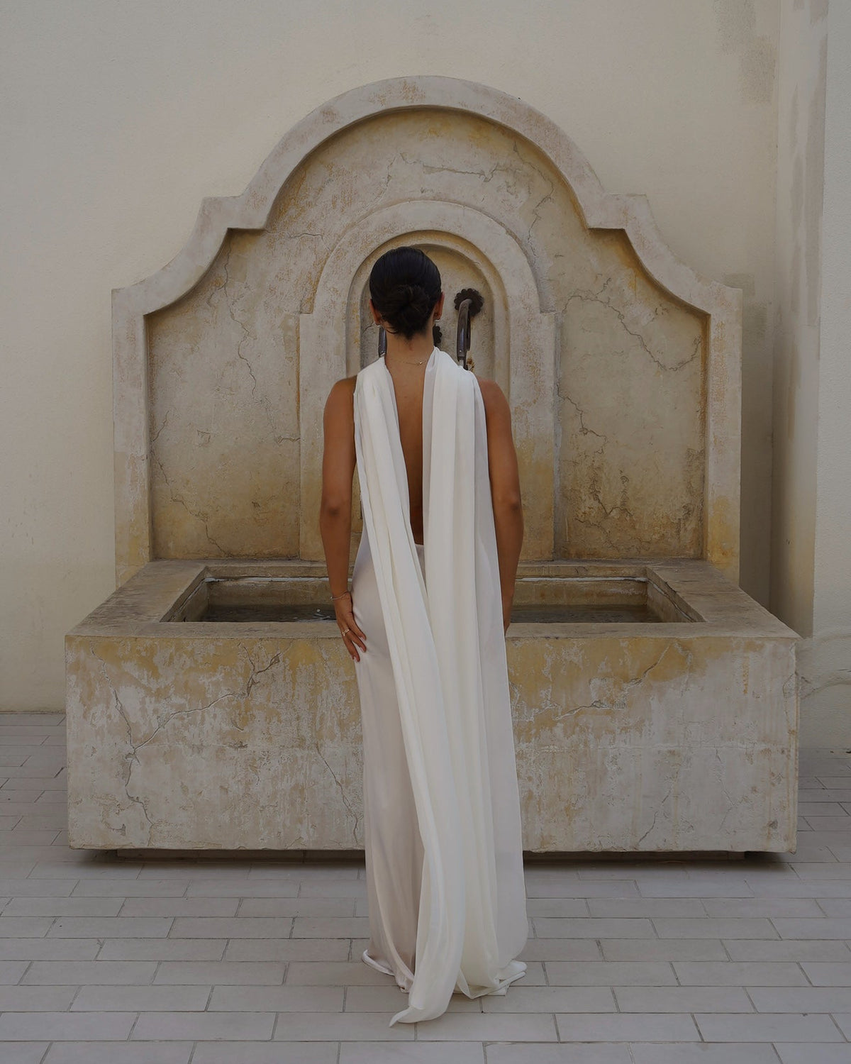A woman stands with her back to the camera in front of a large stone fountain, wearing a long white draped scarf (Farfalla in Ivory by Vedére).