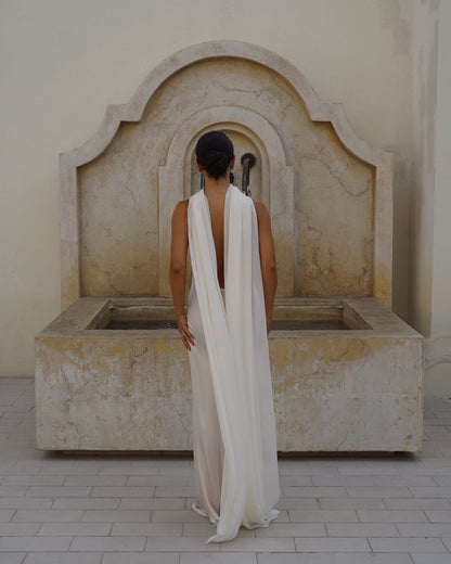 A woman stands with her back to the camera in front of a large stone fountain, wearing a long white draped scarf (Farfalla in Ivory by Vedére).