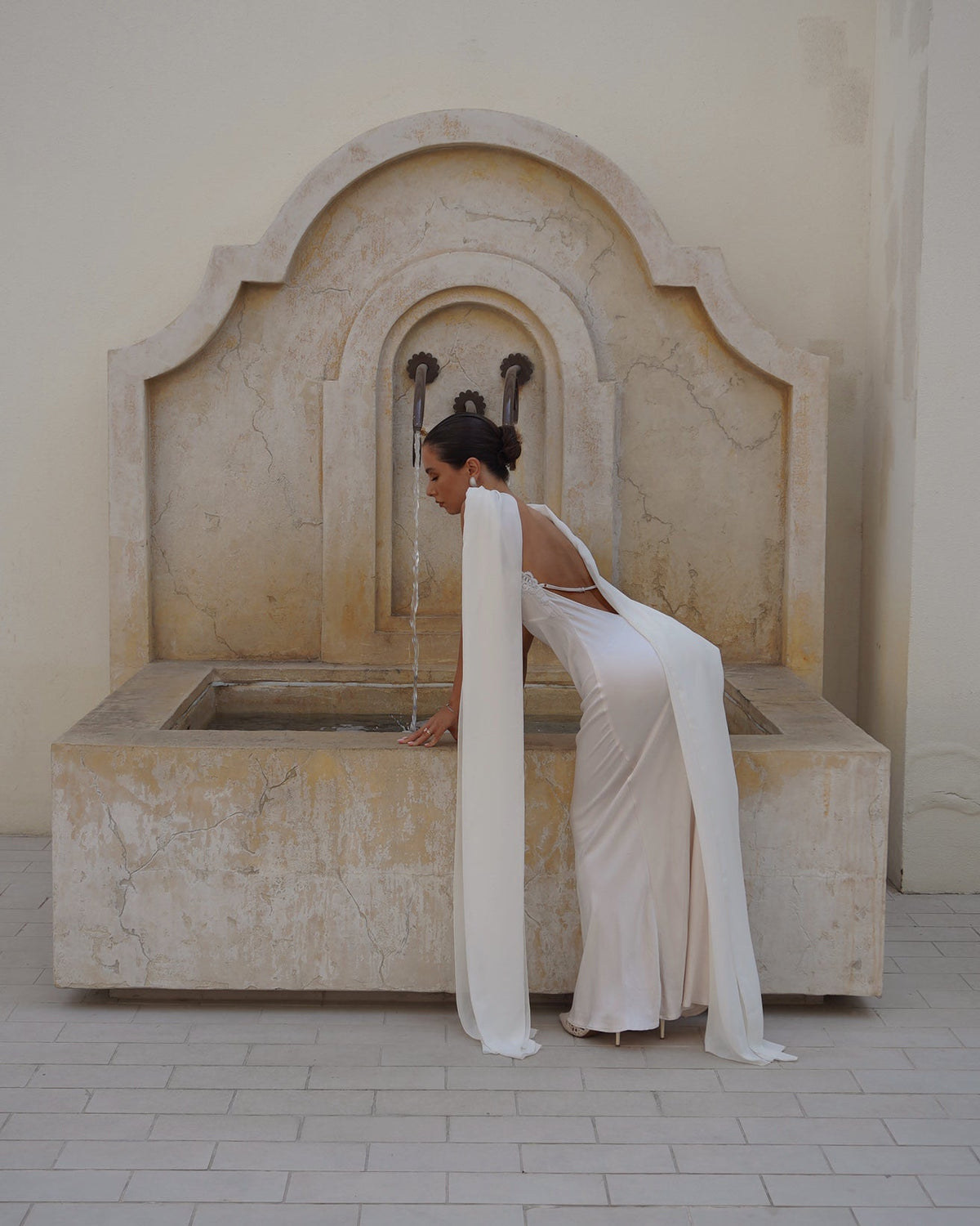 A woman in a flowing white slip wedding gown leans over a large stone fountain in a beige courtyard. She's wearing  the Farfalla in Ivory by Vedére with a low back satin slip dress, and the fountain is set against a plain beige wall.