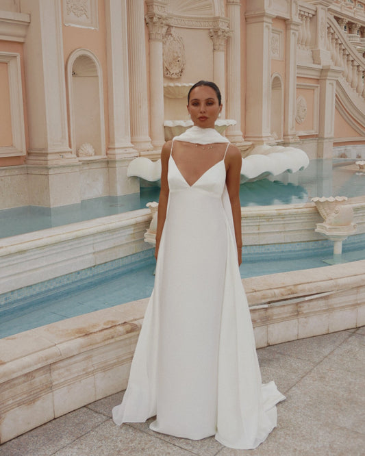 A woman wearing a white sleeveless wedding gown with a deep V-neckline and chiffon scarf stands in front of a grand classical building and blue water feature.