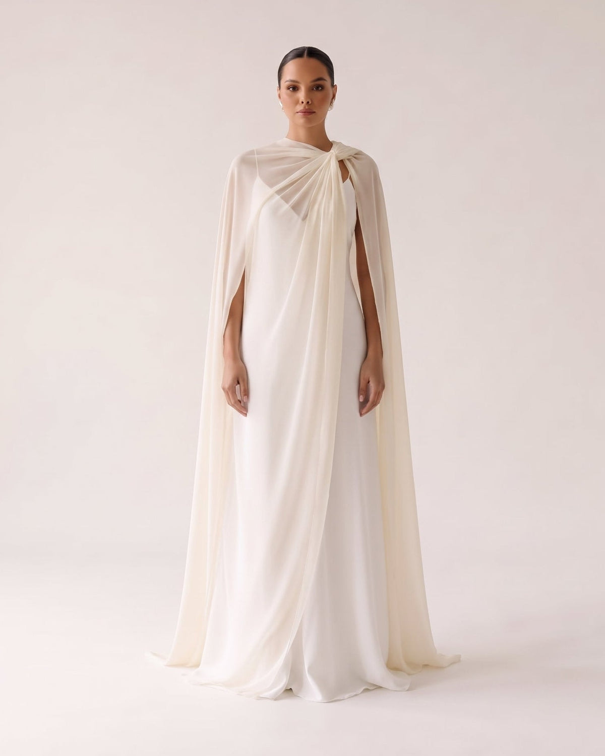 A model stands against a plain light beige studio background wearing a floor-length draped gown with a matching sheer cape. The garment is a warm off-white/ivory tone with a soft, flowing silhouette and no visible branding.