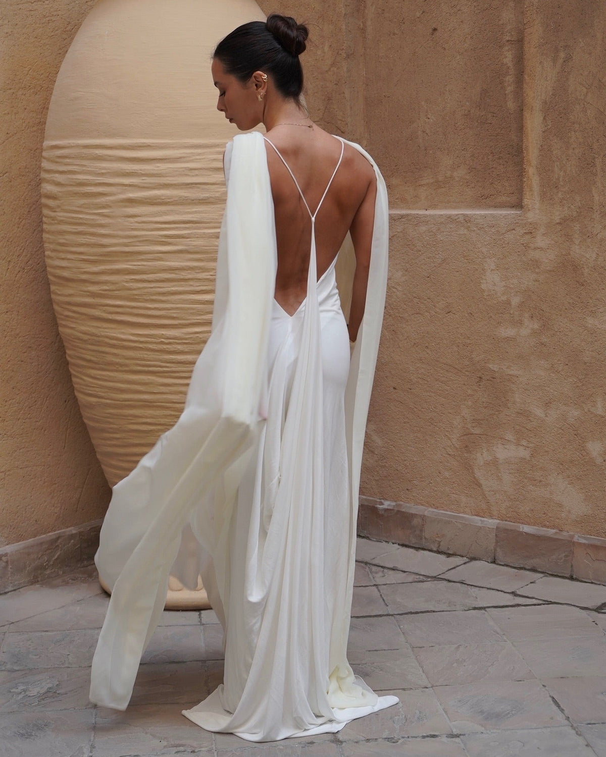 Woman in a white dress with a backless design standing next to a large beige vase against a beige wall.
