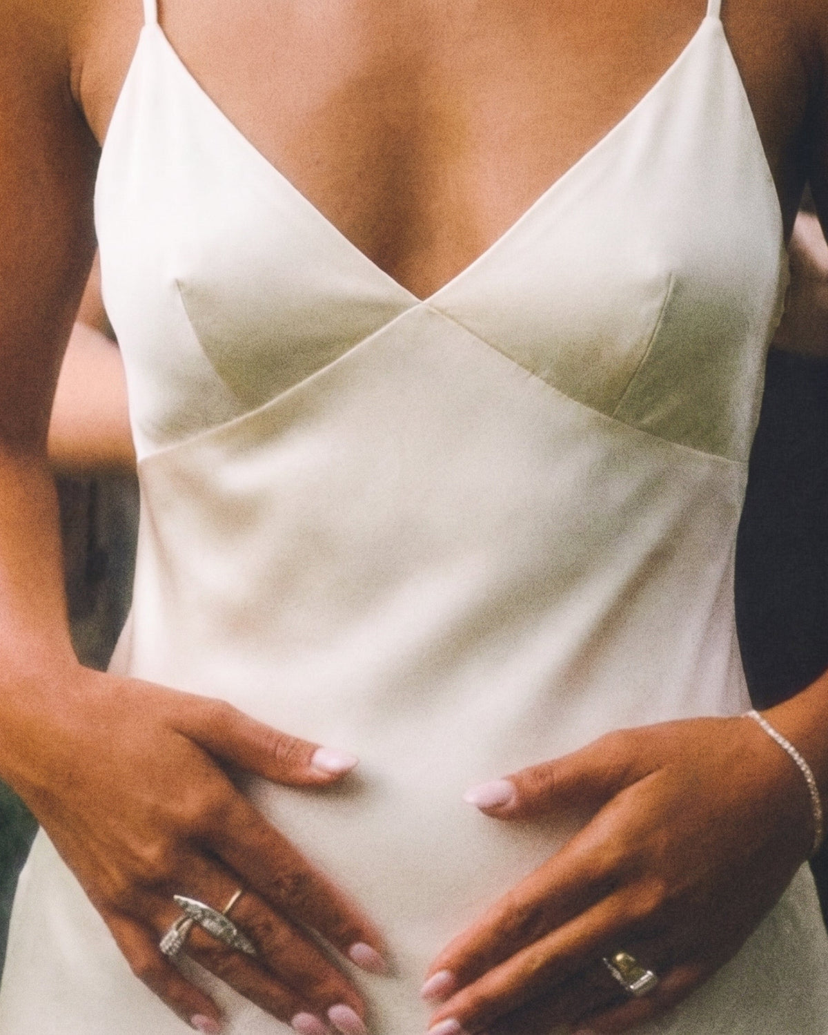 A close-up of a woman wearing a white satin camisole dress, with her hands resting on her belly. She is wearing silver rings and a delicate bracelet.