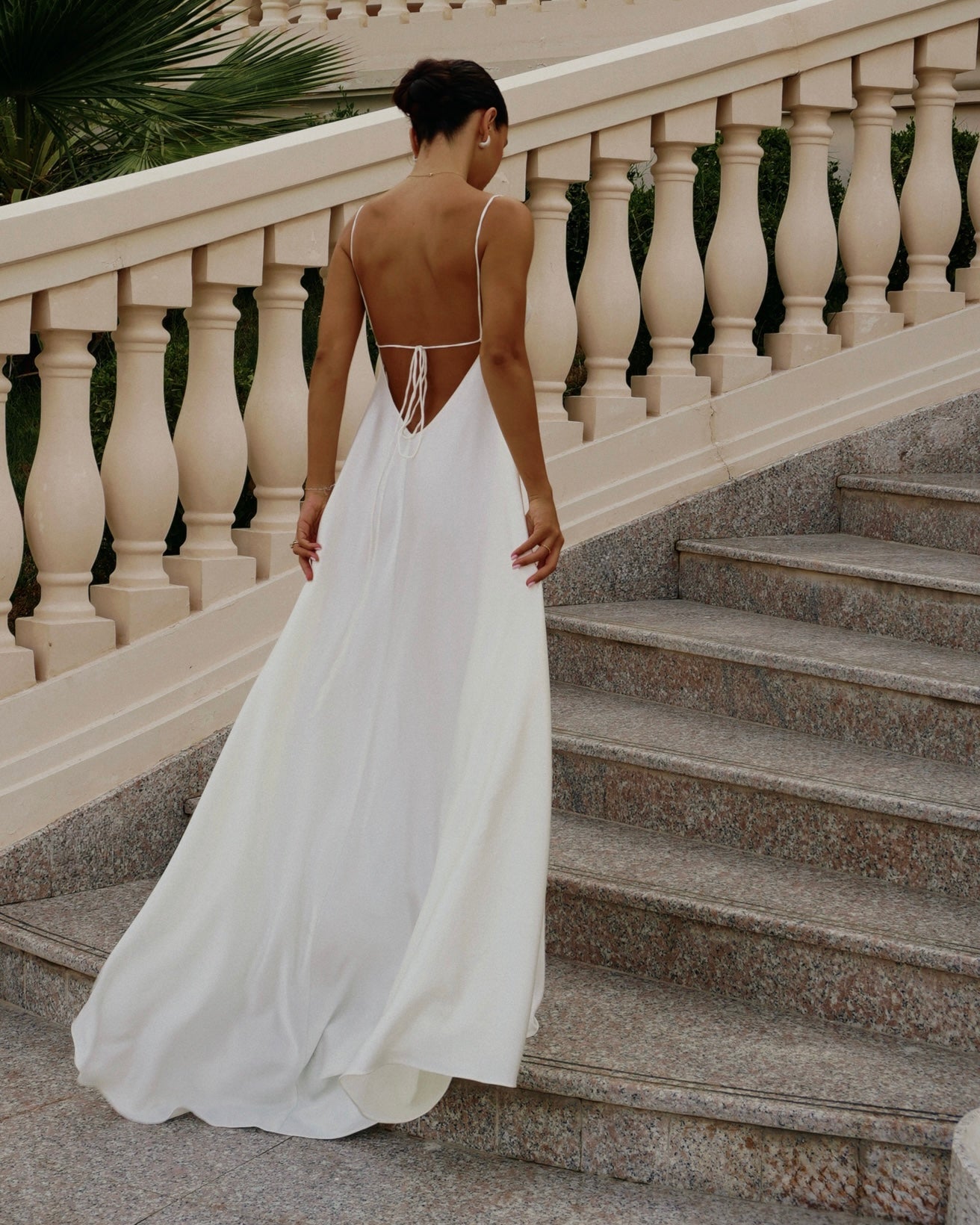 A woman wearing a flowing white sleeveless gown with an open back and thin straps, standing on stone stairs beside a cream-colored balustrade railing.