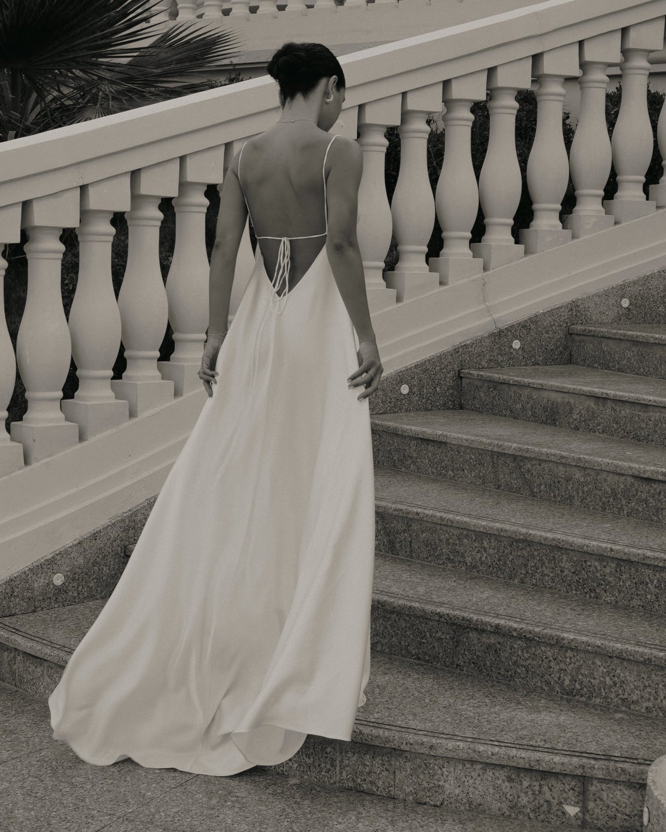 A woman in a flowing white wedding gown stands on an outdoor staircase, holding out the train of the dress. The photo is taken from a high-angle perspective and appears to be in black and white or sepia tones.