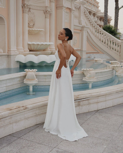 A woman wearing a strapless white floor-length gown with an open back and tie detail, standing in front of a grand beige fountain and staircase.