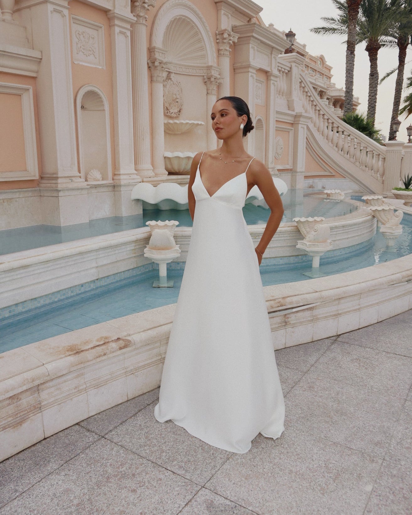 A woman wearing a sleeveless white A-line wedding dress stands in front of a grand classical fountain and terrace with palm trees.