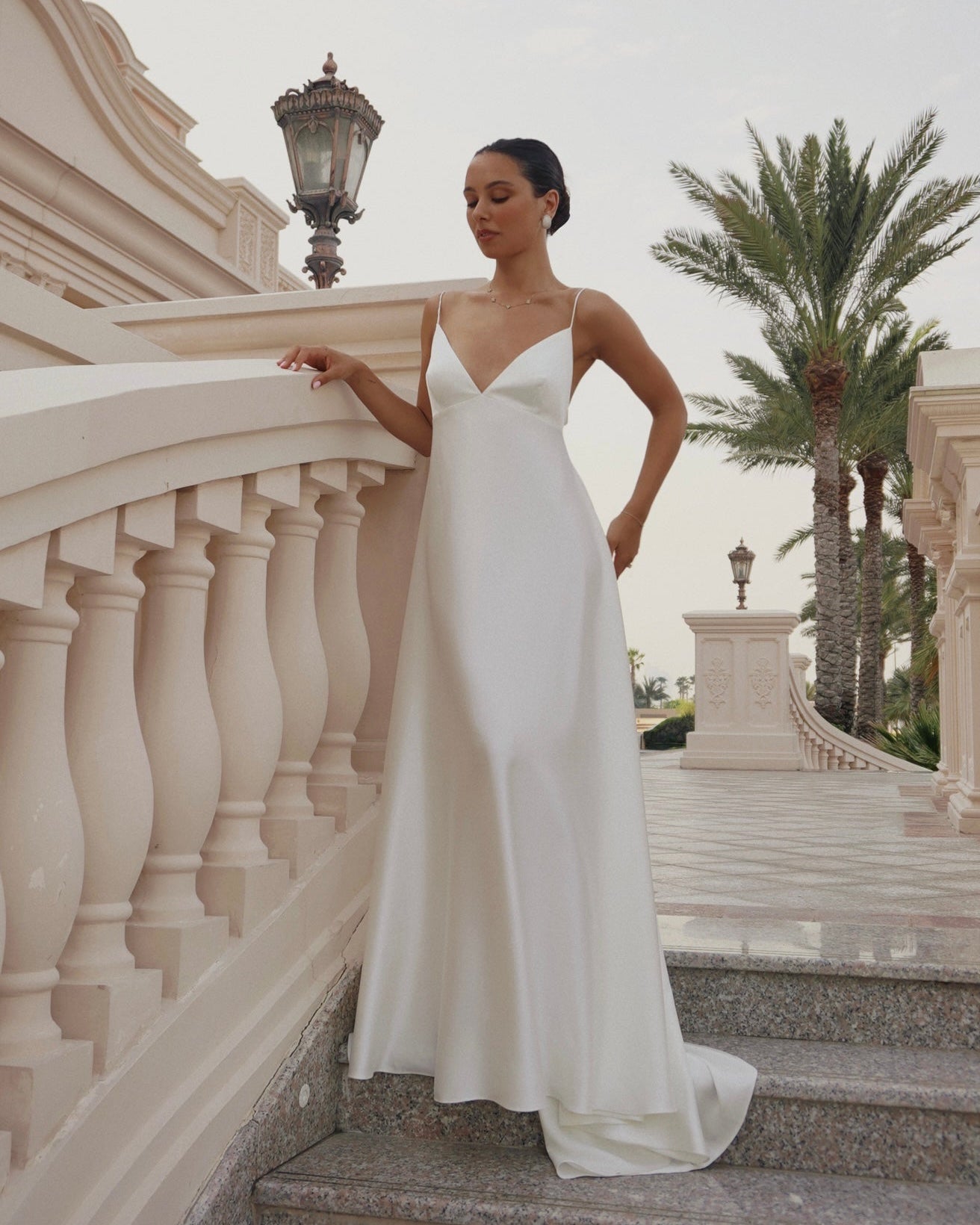A woman wearing a sleeveless white satin wedding gown stands on outdoor stone steps beside a balustrade railing, with palm trees and a pale sky in the background.