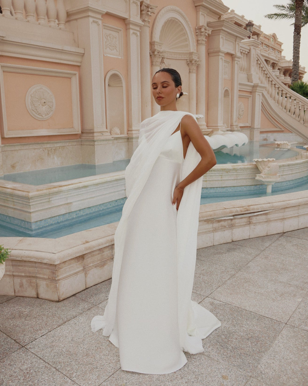 A woman wearing a flowing white bridal gown named the Roma Dress styled with the Vedere Bridal Cape poses outdoors beside a grand beige building and blue water feature.