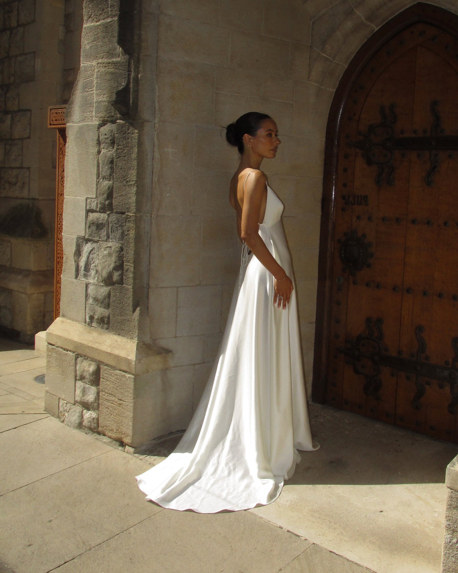 A woman wearing a sleeveless white wedding gown stands in profile beside a large wooden door set within a stone archway.