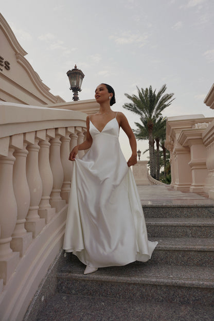 A woman wearing a flowing white wedding gown stands on outdoor stone steps beside a beige building with palm trees in the background.