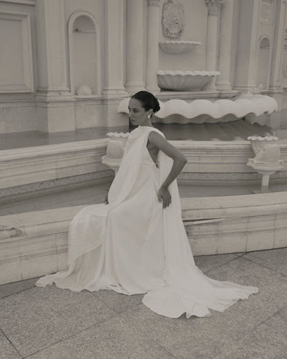 A woman wearing a flowing white bridal gown and bridal cape sits on stone steps in front of a grand classical fountain. The photo is rendered in a soft sepia or monochrome tone, and the background shows ornate architectural details.