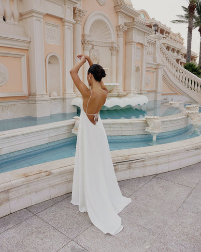 A woman in a flowing white backless gown poses beside a decorative fountain and pale stone building.