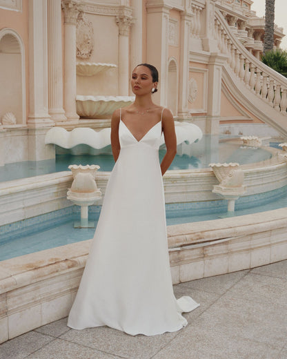 A woman wearing a sleeveless white A-line wedding gown called the Roma Dress with thin straps and a V-neckline, standing in front of a grand beige fountain and staircase.