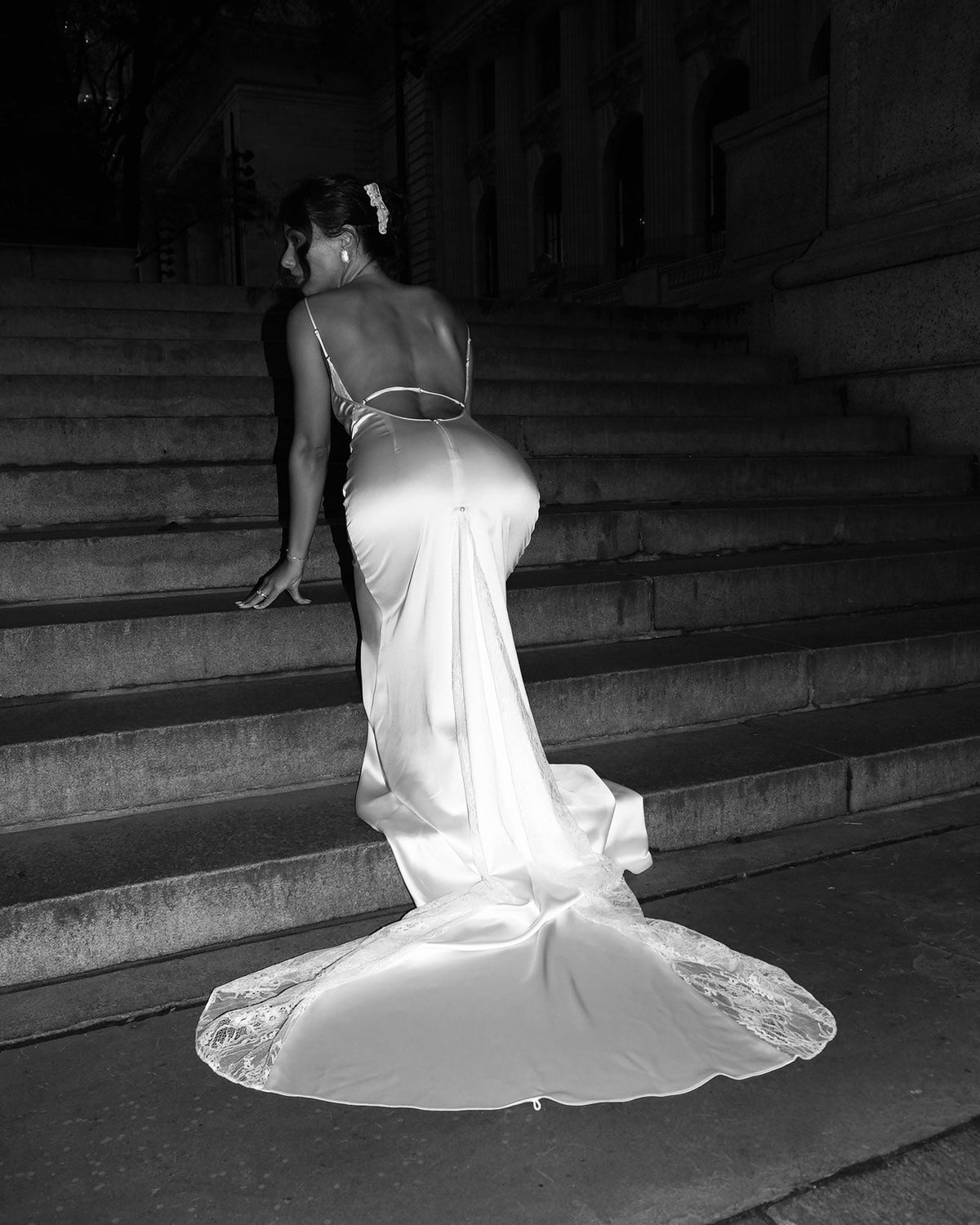 A woman in a white satin wedding gown with lace detailing on the back and hips, photographed from behind as she walks down stone stairs at night.