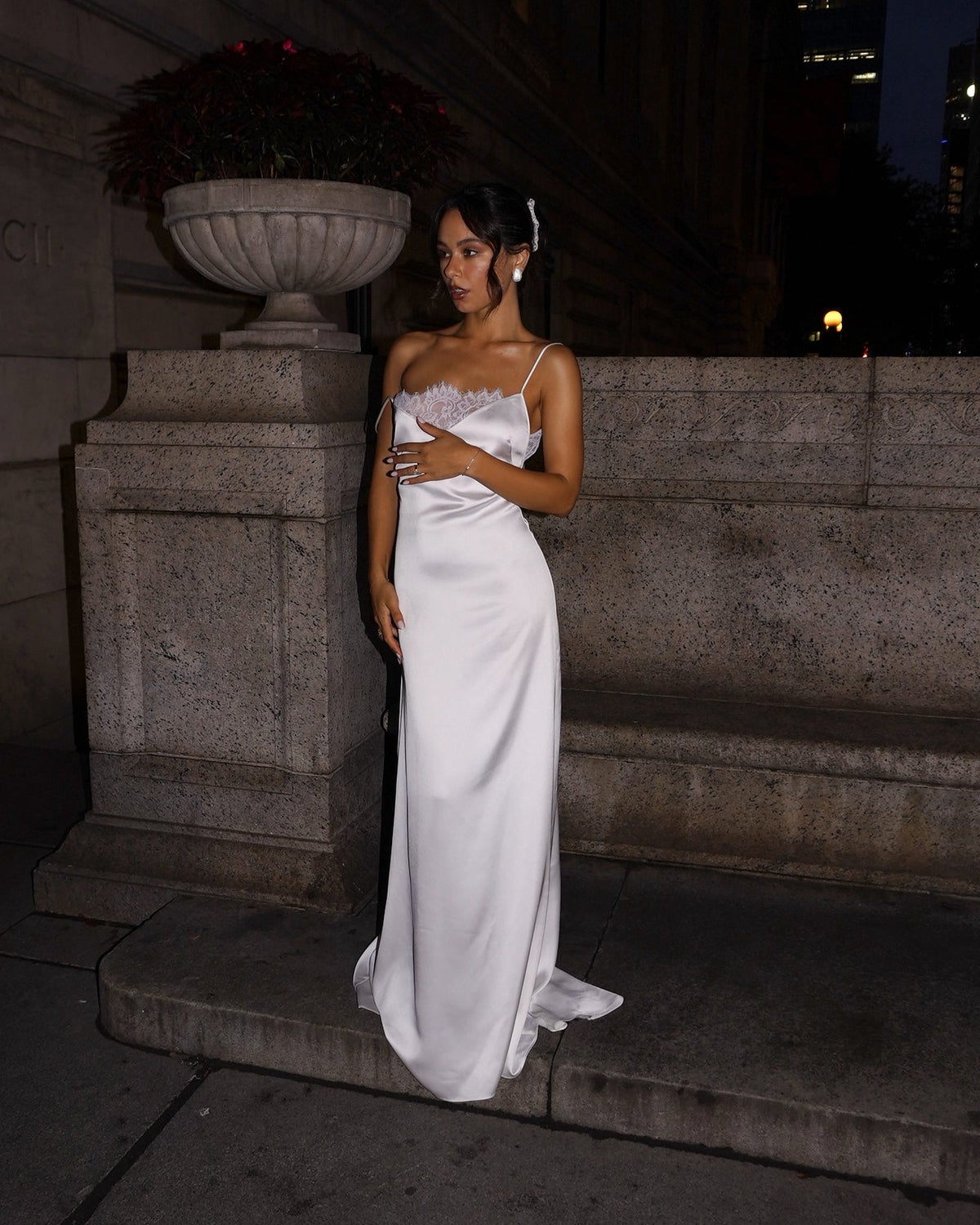 A woman wearing a white satin strapless gown with a sweetheart neckline and lace detailing, standing on stone steps at night.