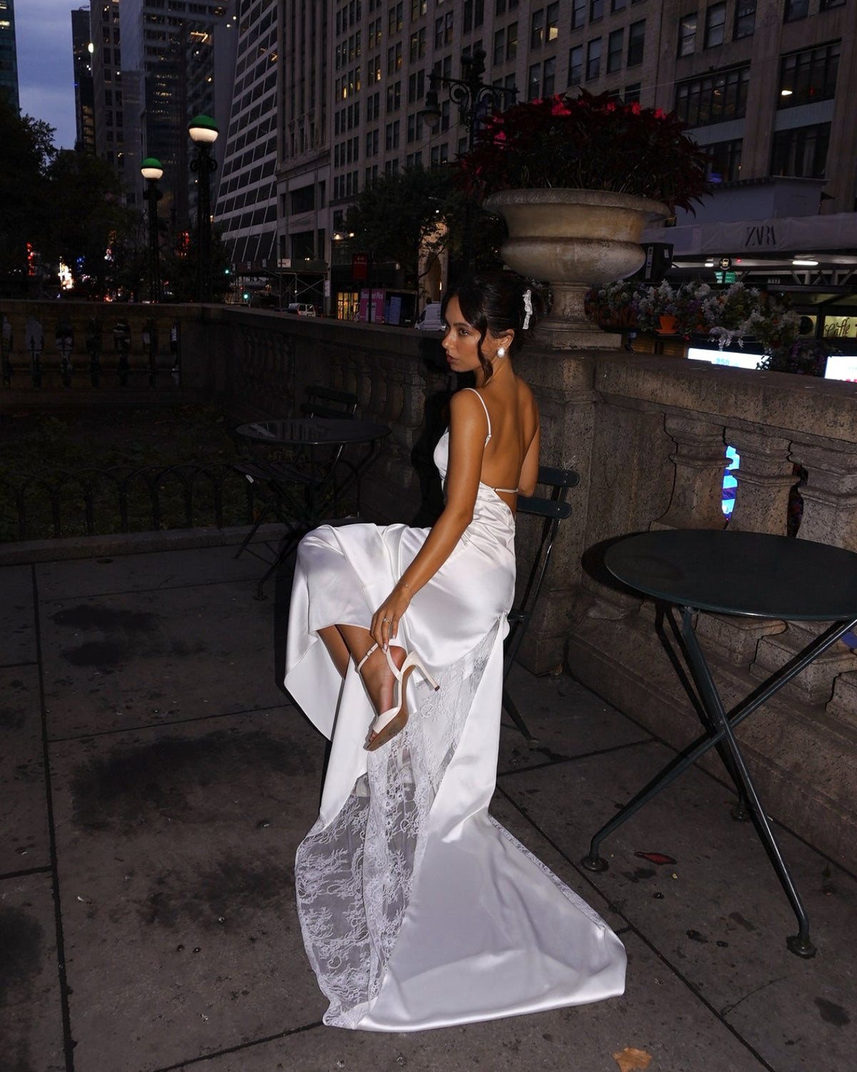 A woman in a white strapless wedding gown with a lace-trimmed hem sits on a stone ledge at dusk, holding her dress out to show the back and train. City buildings, a table, and a planter are visible in the background.