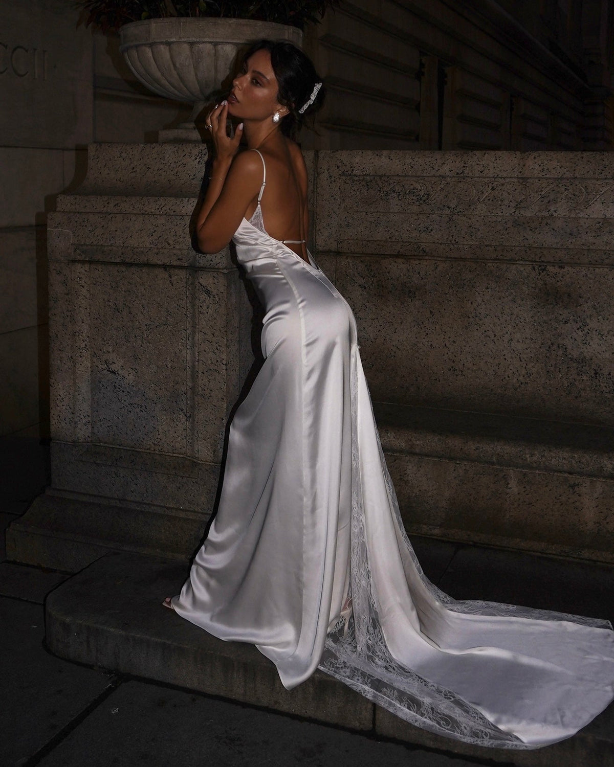 A woman wearing a satin bridal gown with a long train, posing on stone steps at night.