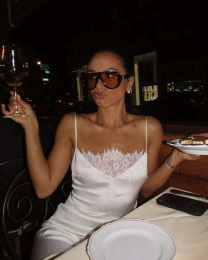 A woman wearing a white spaghetti-strap dress with a lace chest detail, dark sunglasses, and a bracelet, posing at a restaurant table with a wine glass and plates.