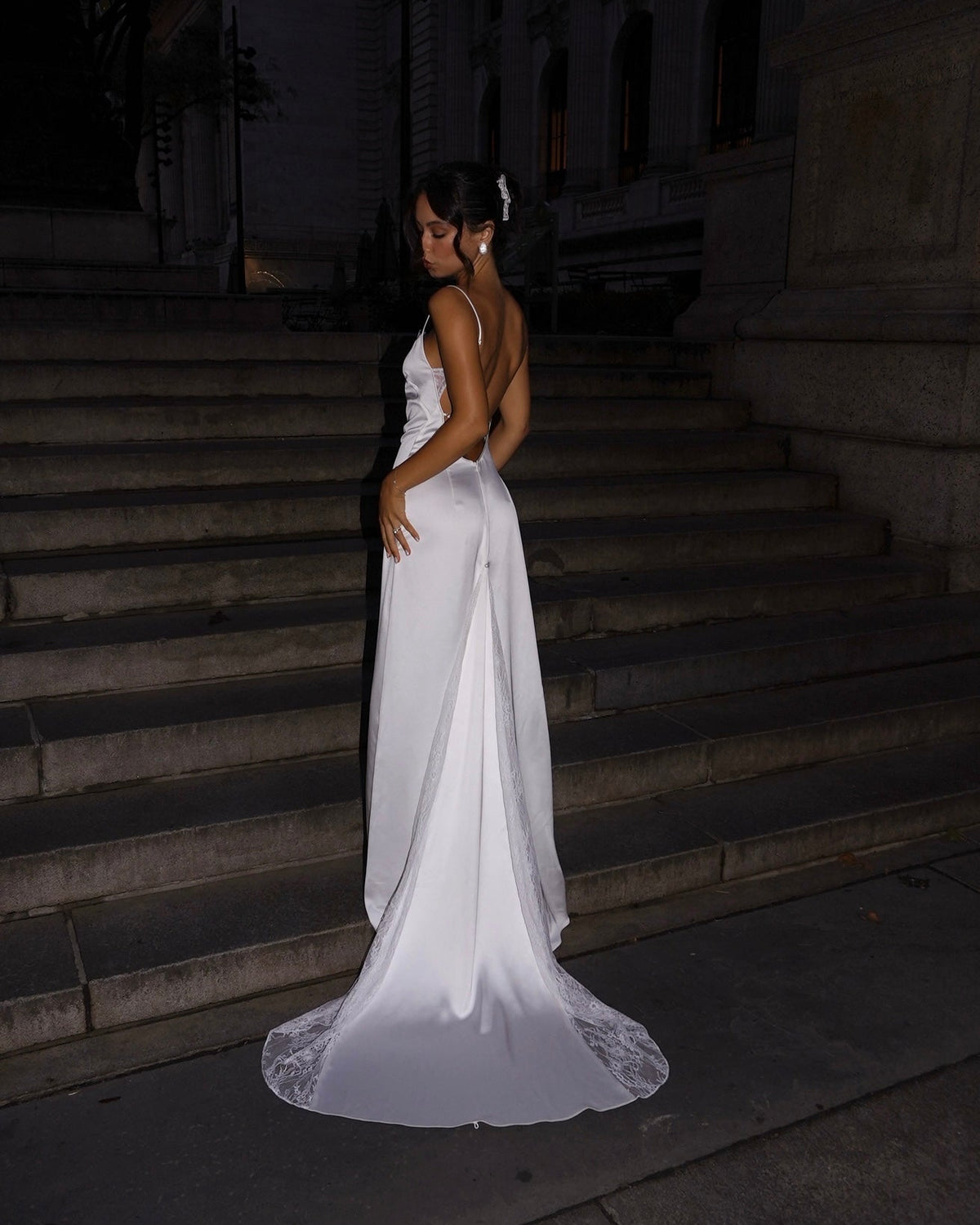 A woman wearing a sleeveless white wedding gown with a fitted bodice, draped satin skirt, and lace-trimmed hem stands on stone steps at night, looking back over her shoulder.
