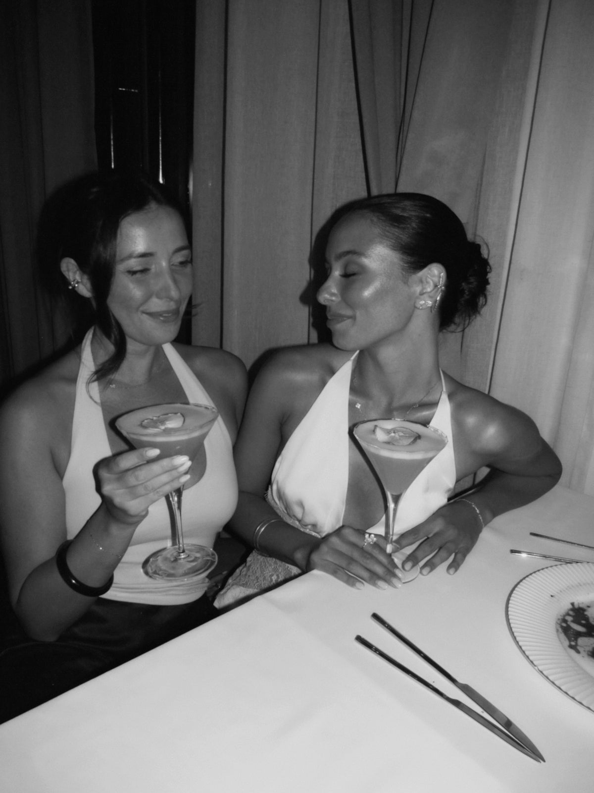 Black and white photo of two Vedére founders, Isabella and Rachael, sitting at a table, each holding a martini glass. They are wearing white halter neck dresses.